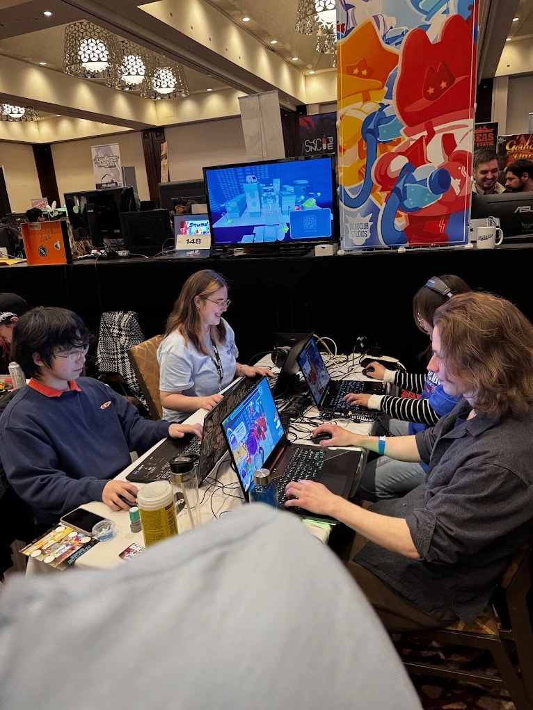The team demoing the game live on laptops for attendees at a showcase booth, with the Deadclub
                Studios banner visible behind them.